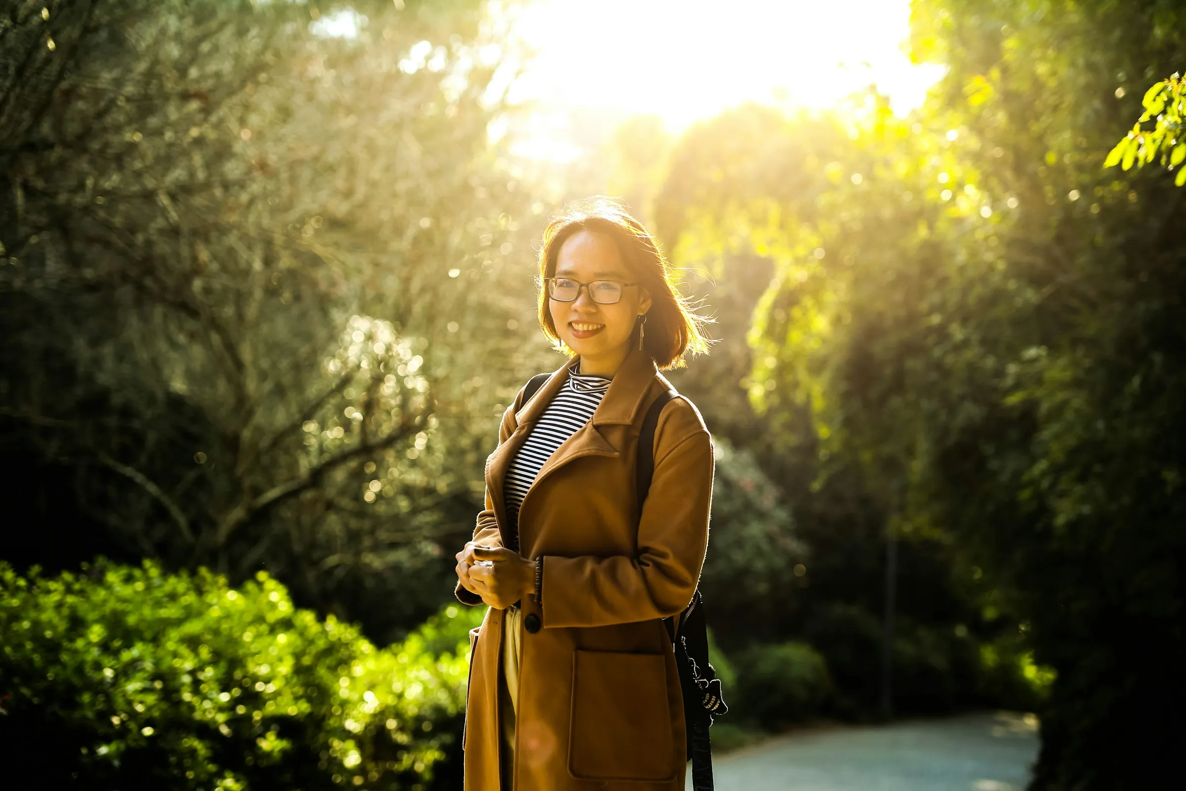A woman standing outdoors on a tree-lined path in morning sunlight
